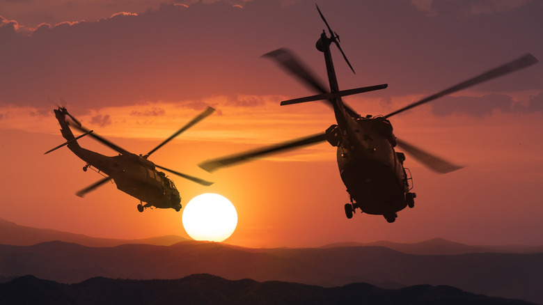 A silhouette aerial view of Army helicopters flying over mountains at sunset.
